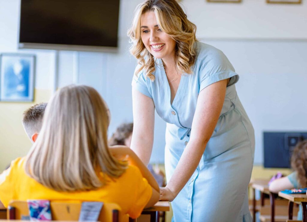 Smiling teacher assisting young girl with assignment at school desk in classroom.
