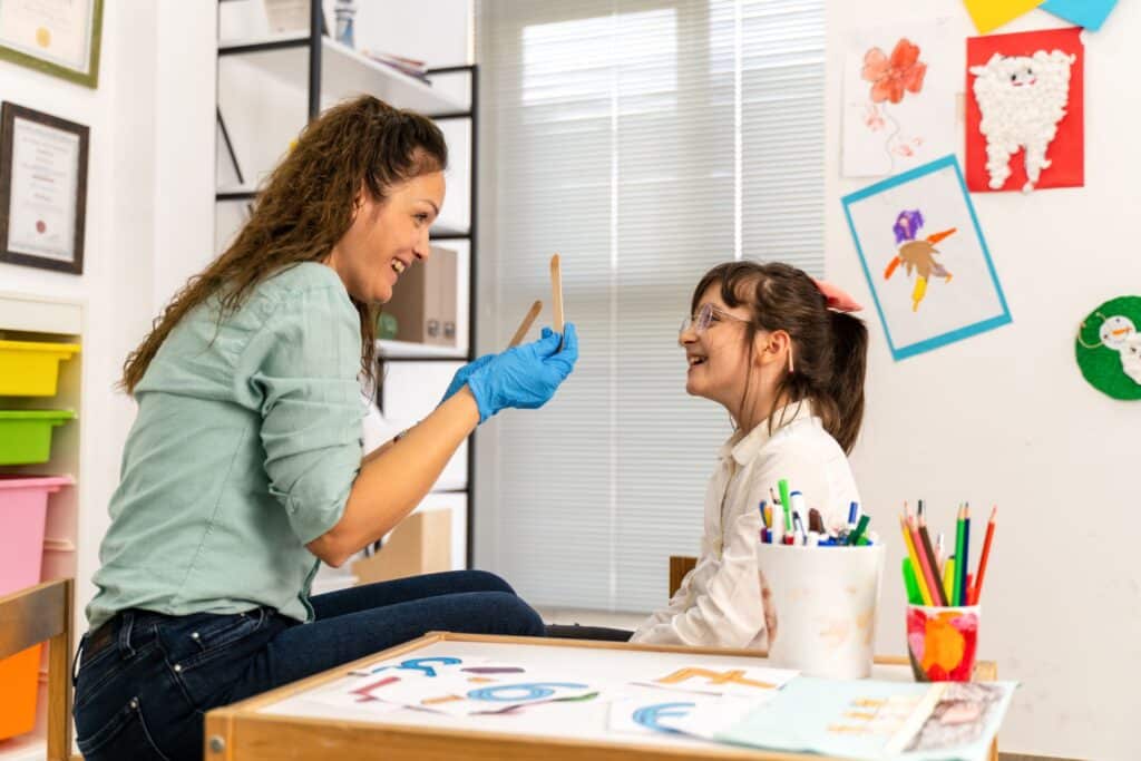 Woman holding up two tongue depressors while a little girl giggles in front of her