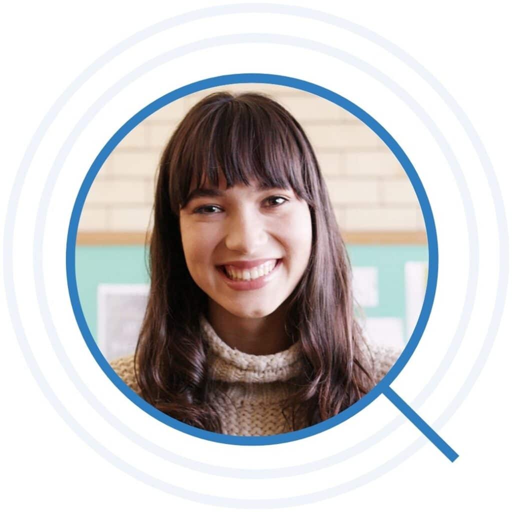 Brunette teacher with bangs smiling in front of a bulletin board
