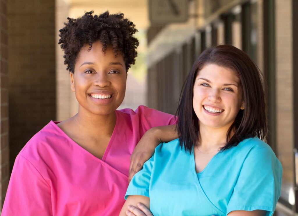 Two smiling healthcare professionals in colorful scrubs standing together outside a medical facility.