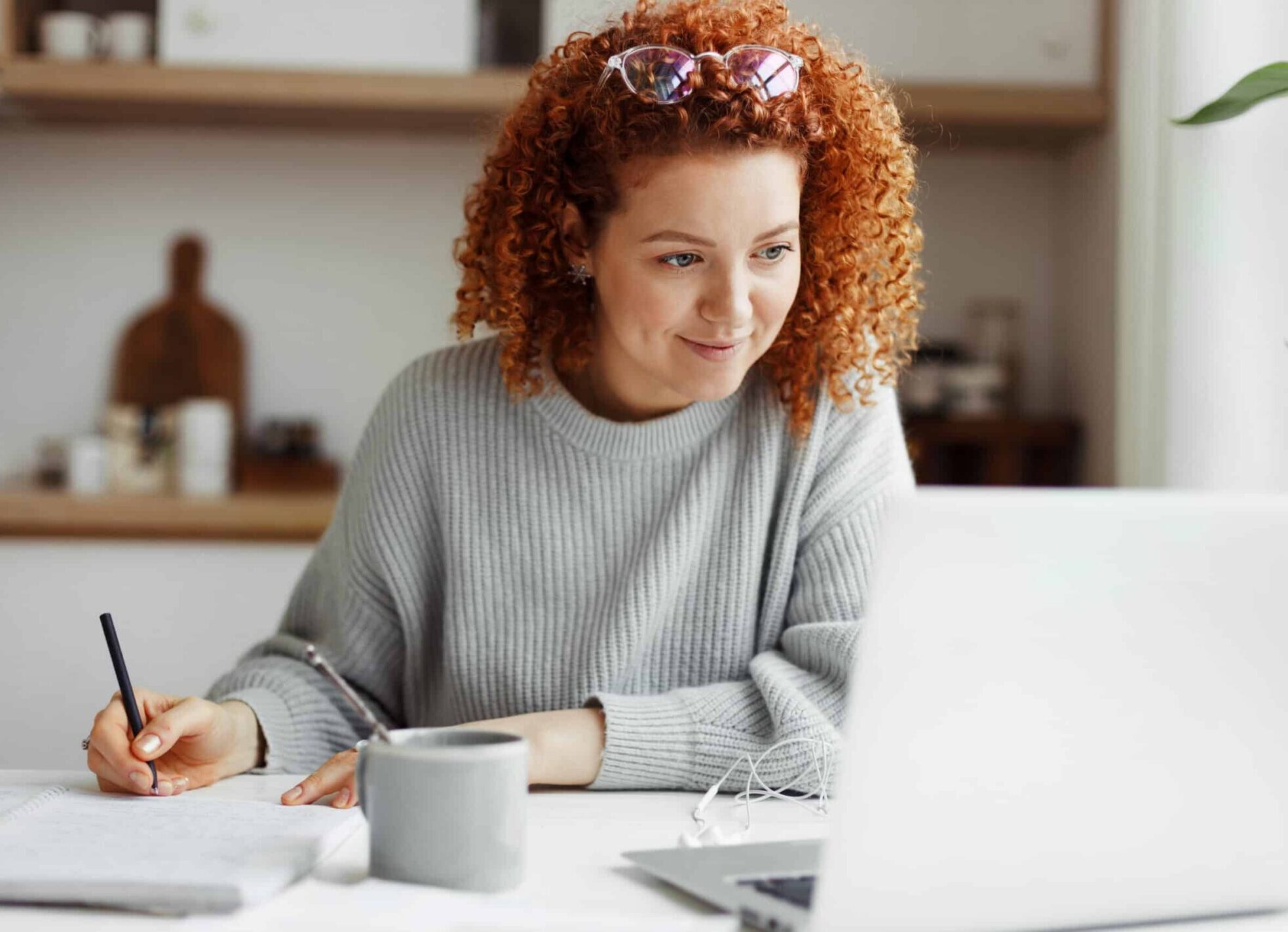Portrait of happy woman participating in a virtual interview sitting at her kitchen table, taking notes, and looking at the screen with interest.