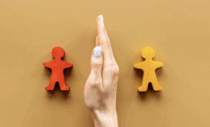 A hand positioned vertically like a barrier separates two wooden human-shaped figures — one painted red and the other yellow — set against a tan background, symbolizing conflict or division between two people.