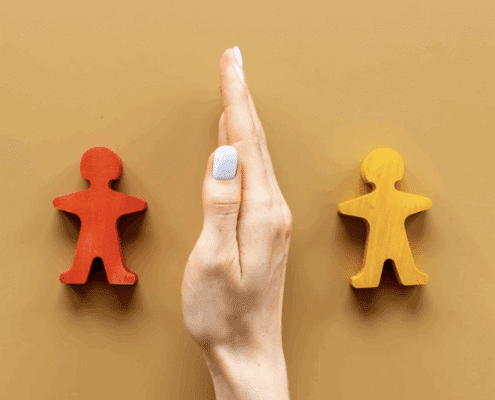 A hand positioned vertically like a barrier separates two wooden human-shaped figures — one painted red and the other yellow — set against a tan background, symbolizing conflict or division between two people.