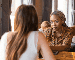 Two women sit across from each other at a table, engaged in a serious conversation.