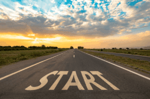 An open road stretching into the distance under a colorful sunrise or sunset sky, with the word 'START' painted in large white letters on the asphalt.