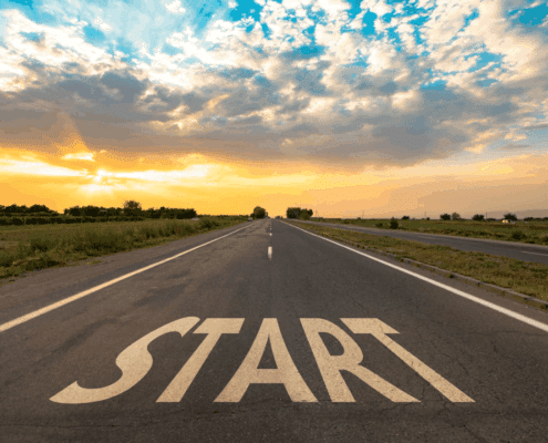 An open road stretching into the distance under a colorful sunrise or sunset sky, with the word 'START' painted in large white letters on the asphalt.