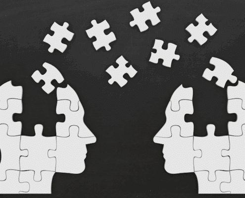 Two human heads made of white puzzle pieces facing each other on a black background, with several puzzle pieces floating between them, symbolizing knowledge transfer, communication, or shared understanding.