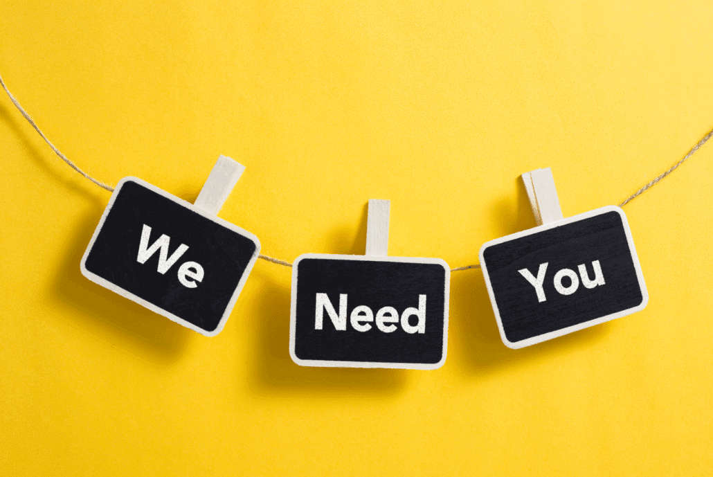 Three small black signs with white borders clipped to a string against a bright yellow background. The signs display the words 'We Need You' in white letters.