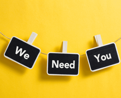 Three small black signs with white borders clipped to a string against a bright yellow background. The signs display the words 'We Need You' in white letters.